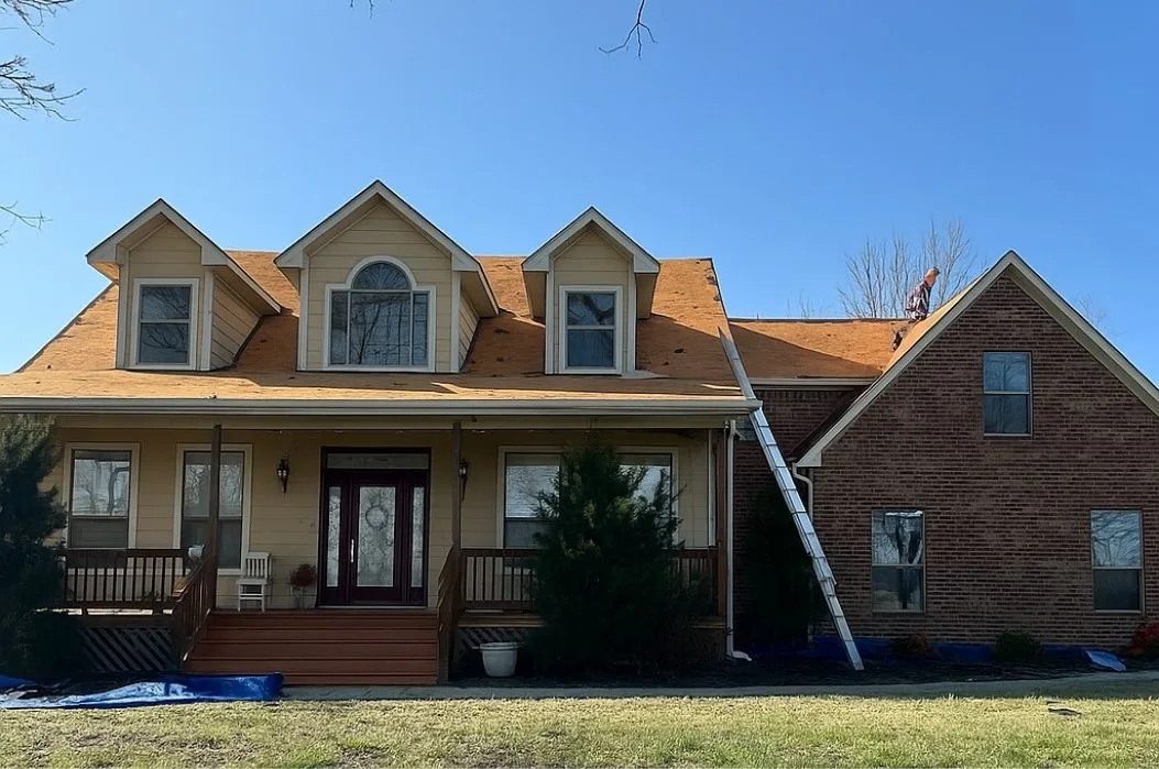 Roof stripped to fresh decking with a crew member working the ridge and tarps protecting the landscaping below
