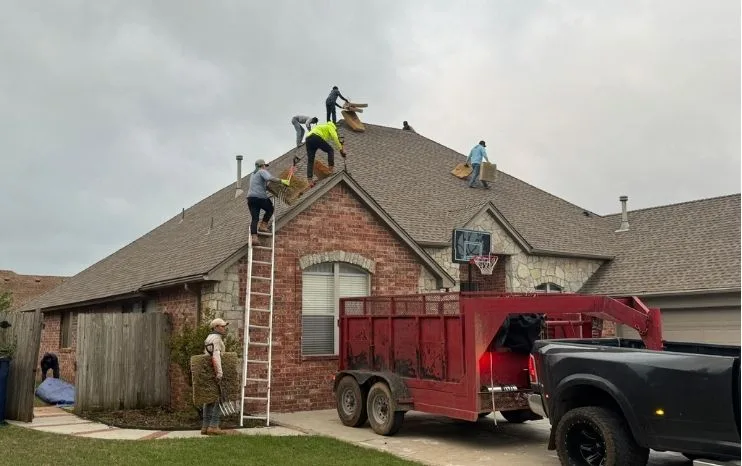 Shoemaker Snoddy crew tearing off shingles on a brick Oklahoma home with the company truck and dump trailer in the driveway